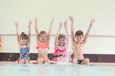Die Teilnehmer des Kinder-Schwimmkurses sitzen am Beckenrand und heben die Arme in die Luft.