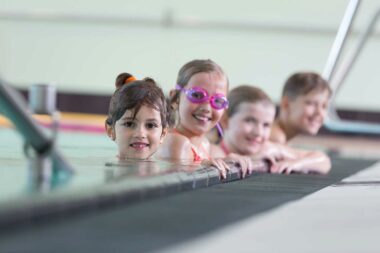 Die Teilnehmer des Kinder-Schwimmkurses halten sich am Beckenrand fest und lächeln.