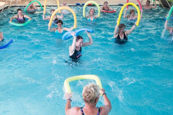 Die Trainerin macht eine Übung mit Poolnudel im Wasser vor, die Teilnehmer machen ihr nach.